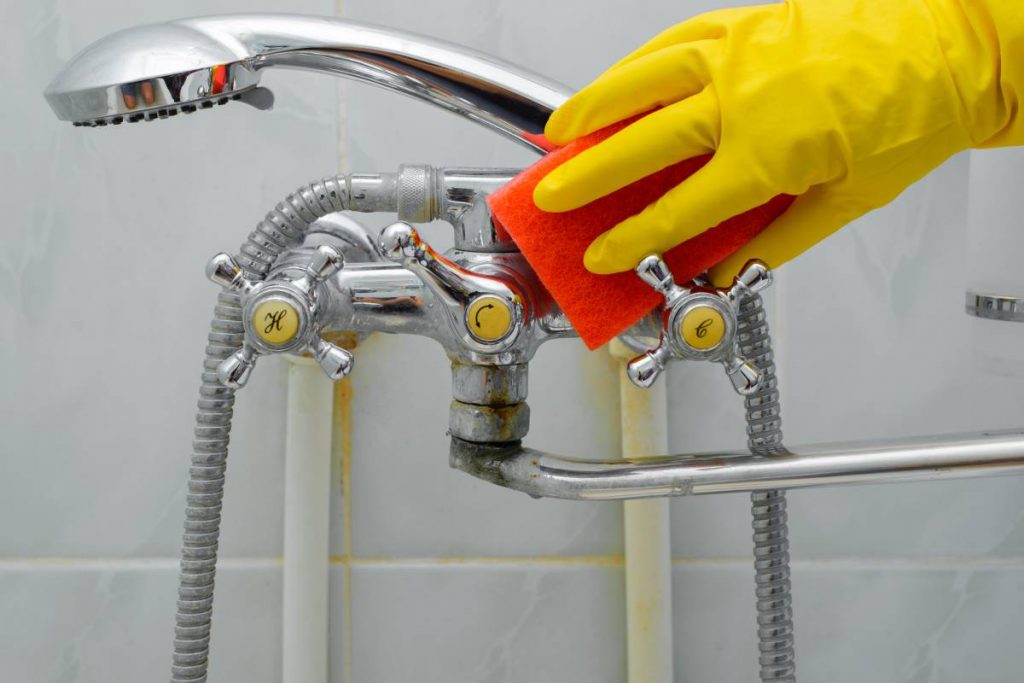 Close-up of hand in rubber glove washing dirty, rusty faucet with shower, cleaning bathroom.